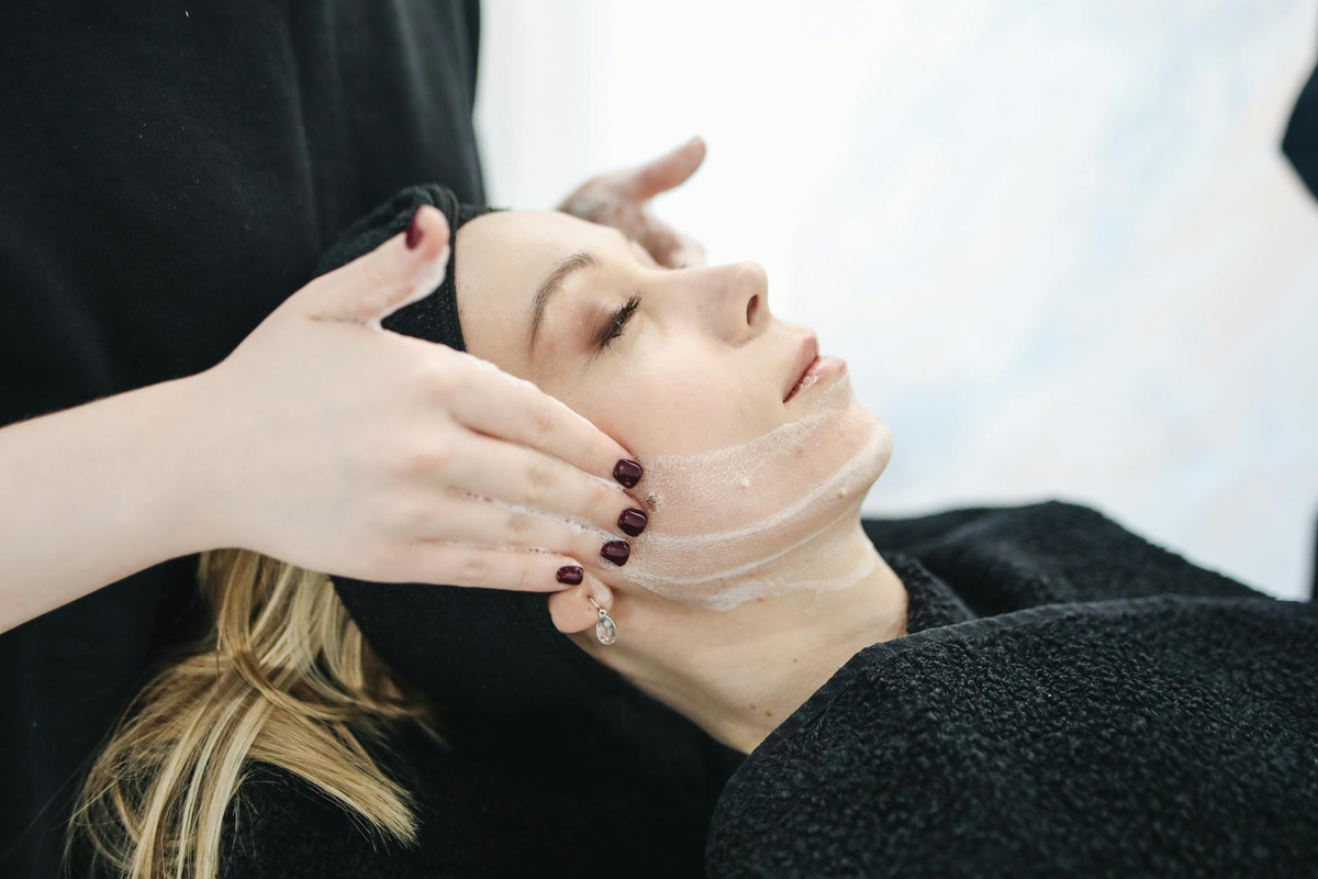 Hands apply a foamy cleanser to a woman's face during a relaxing spa facial, representing advanced skin care for a flawless complexion.