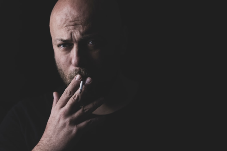 A contemplative bald man with a beard holds a lit cigarette to his lips, casting deep shadows across his face against a dark background.