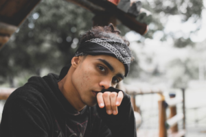 A pensive young man in a black bandana and hoodie looks intently forward, hand thoughtfully positioned at his mouth, symbolizing deep concentration and focus in an outdoor setting.