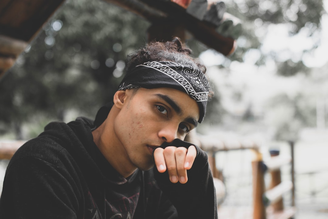 A pensive young man in a black bandana and hoodie looks intently forward, hand thoughtfully positioned at his mouth, symbolizing deep concentration and focus in an outdoor setting.