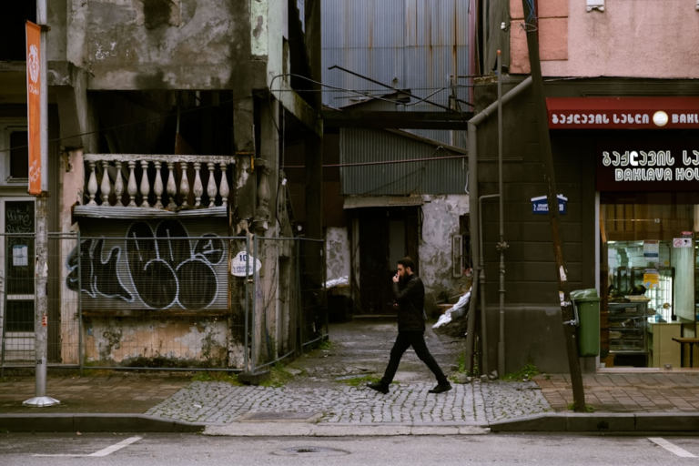 Gritty urban scene featuring a man walking along a cobblestone street lined with crumbling architecture, a graffiti-covered storefront, and a Baklava House in Georgia.