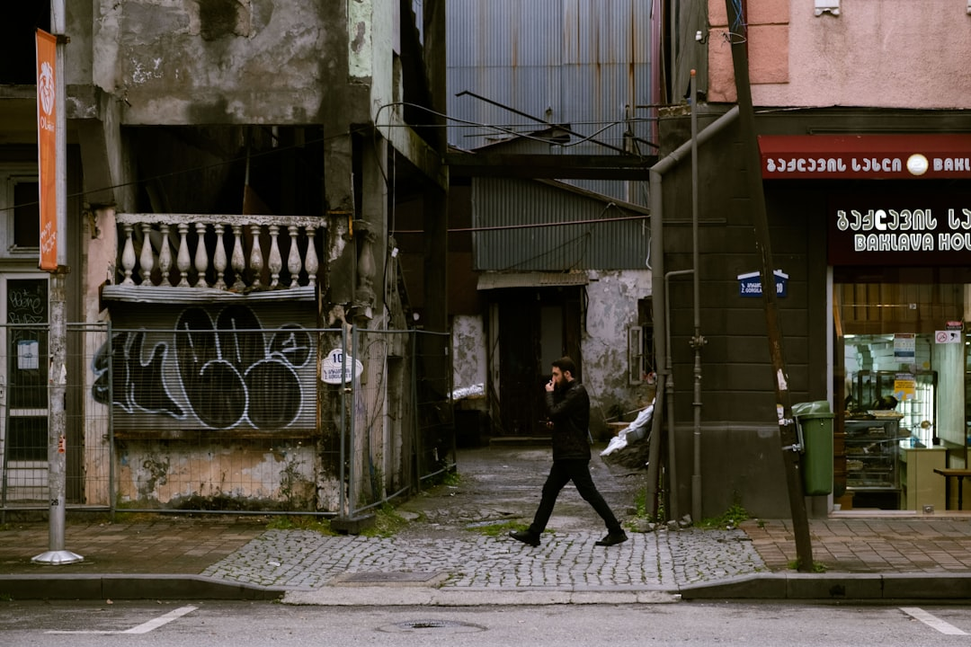 Gritty urban scene featuring a man walking along a cobblestone street lined with crumbling architecture, a graffiti-covered storefront, and a Baklava House in Georgia.