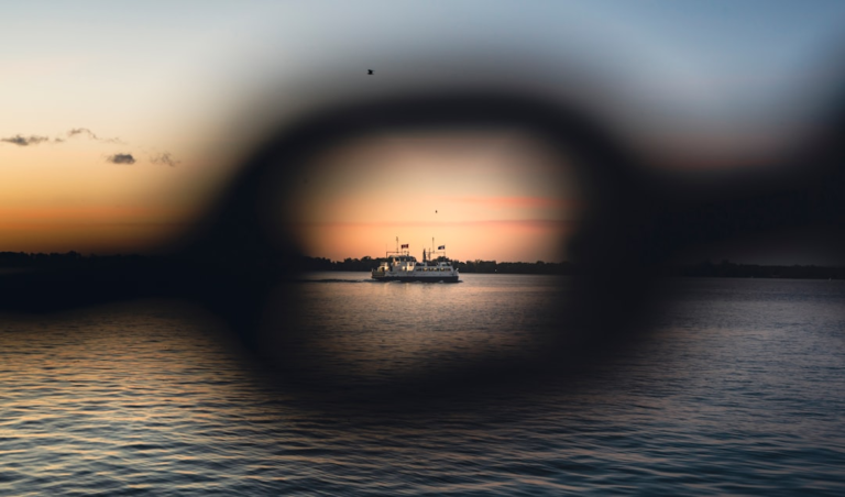 A ferry boat sails clearly in the focused center of a sunset over water, while the surrounding visual field is blurred and dark, simulating central vision loss often experienced with macular degeneration.