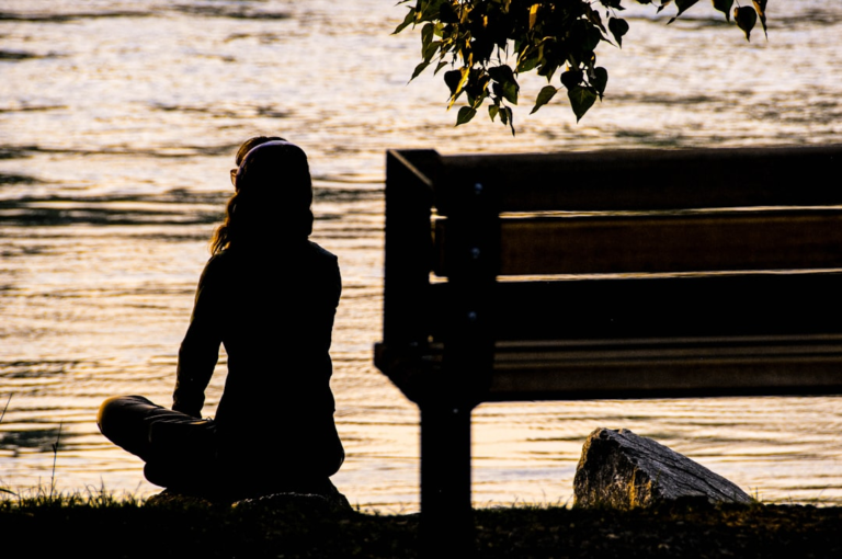 Silhouette of an individual in a cross-legged meditative pose by shimmering golden water at dusk, representing calm focus for advanced breathwork and mental regulation.