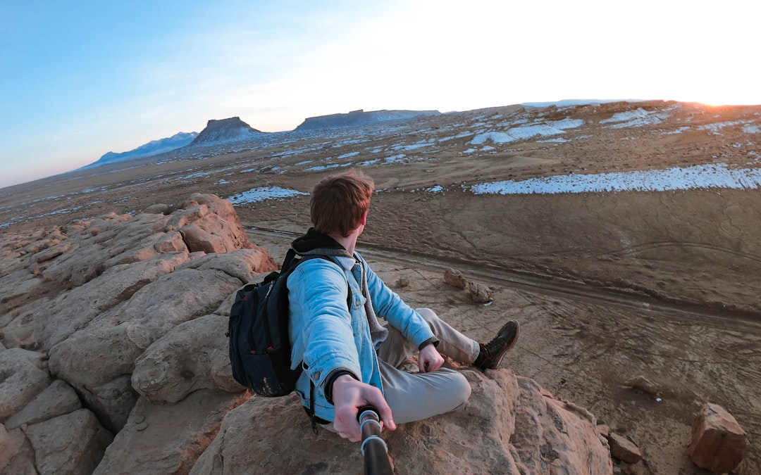 Young man with a backpack sitting on a rugged cliff, gazing over a vast, sunlit landscape with distant mesas and snow patches, embodying peak vitality and a sense of freedom.