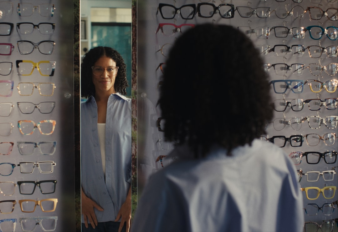 A smiling woman with curly hair tries on a pair of stylish glasses in an optical store mirror, surrounded by a diverse selection of frames suitable for blue light blocking.