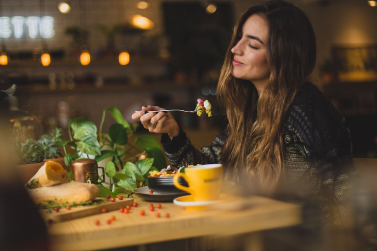 A content young woman with long hair savors a forkful of colorful, nutritious food at a cozy restaurant table, emphasizing mindful healthy eating for optimal eye health.
