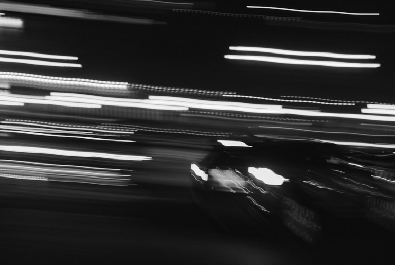 Black and white long exposure shows a blurred car with bright headlights and streaking light trails on a dark night road, emphasizing enhanced night vision.