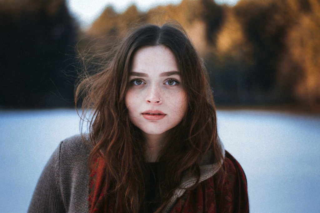 Young woman with clear, radiant skin and voluminous brown hair looks directly ahead, her natural beauty highlighted against a soft outdoor backdrop, embodying ageless vitality.