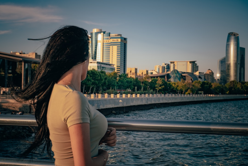 Woman with windswept dark hair gazes contemplatively at a modern city skyline across a sunlit bay, reflecting on her beauty journey amidst urban serenity.