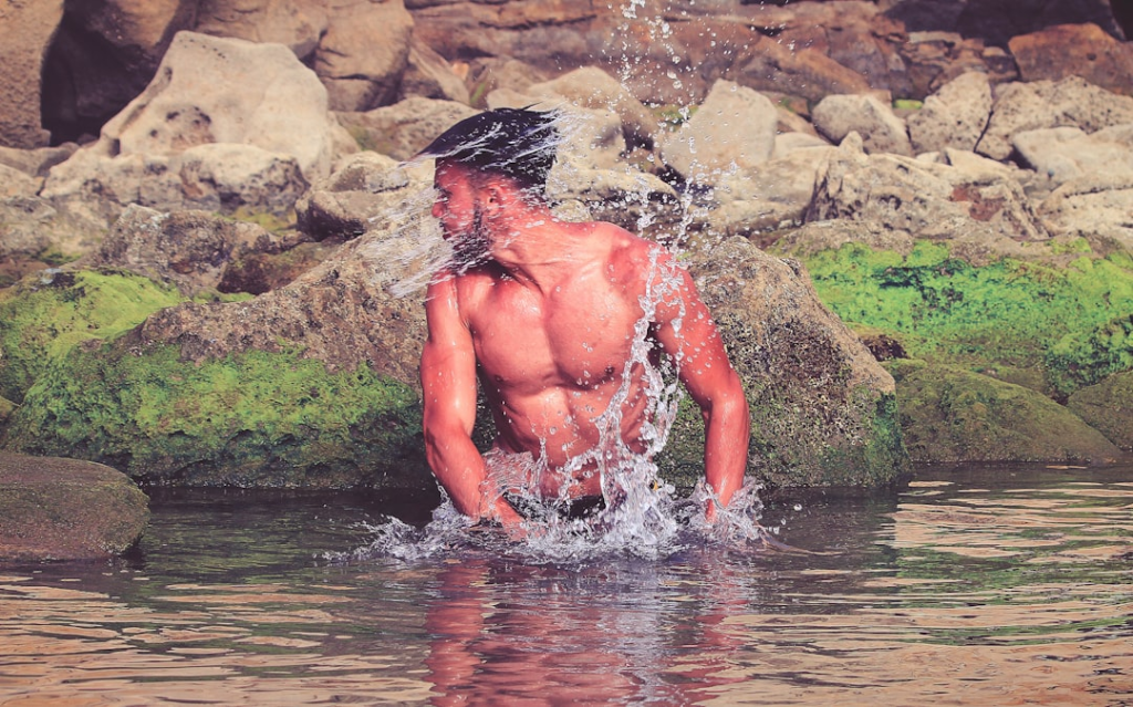Muscular man vigorously splashes water in a natural pool surrounded by moss-covered rocks, embodying renewed vitality and optimized energy.