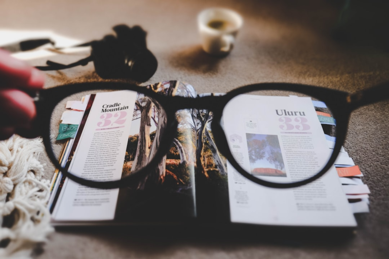 Clear vision through a pair of eyeglasses frames an open travel magazine featuring destinations like Cradle Mountain and Uluru, emphasizing the importance of sharp eyesight and proactive macular health for AMD prevention.