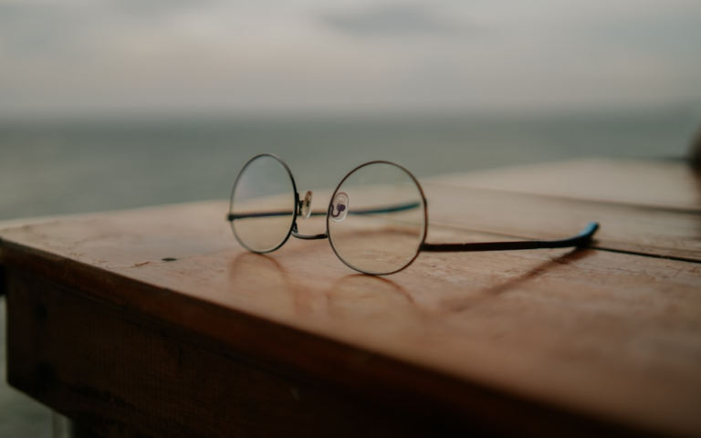 Round reading glasses rest on a rustic wooden table overlooking a blurred ocean horizon, symbolizing a focus on vision and eye health in the context of Wet AMD support.