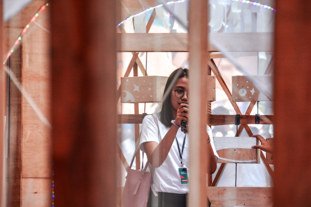 A focused young woman in glasses holds a phone through a warm wooden structure decorated with celestial designs, capturing a moment of active observation or mental engagement.