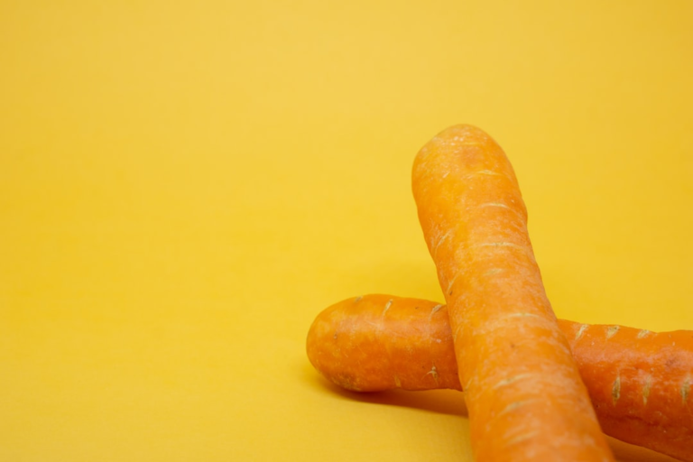 Two vibrant orange carrots laid diagonally on a bright yellow background, representing natural nutrition crucial for optimal vision and eye health.