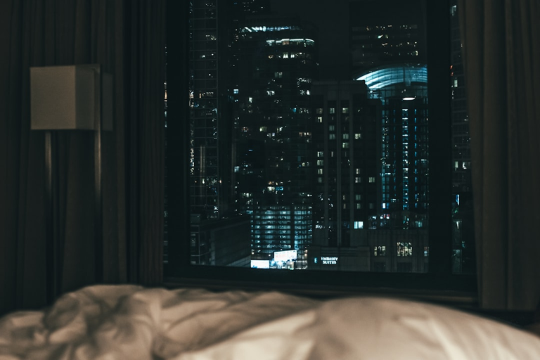 A dark bedroom overlooks a vibrant city skyline at night, filled with brightly lit buildings, underscoring the prevalence of artificial light exposure impacting sleep and circadian rhythm.