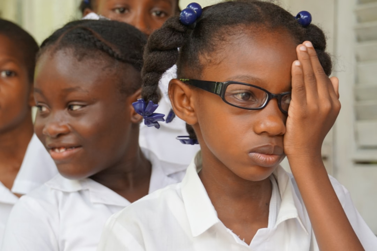 A young girl wearing black-rimmed glasses covers one eye during a vision test, surrounded by other children in a soft-focus setting, emphasizing the importance of eye health screenings for conditions such as astigmatism.