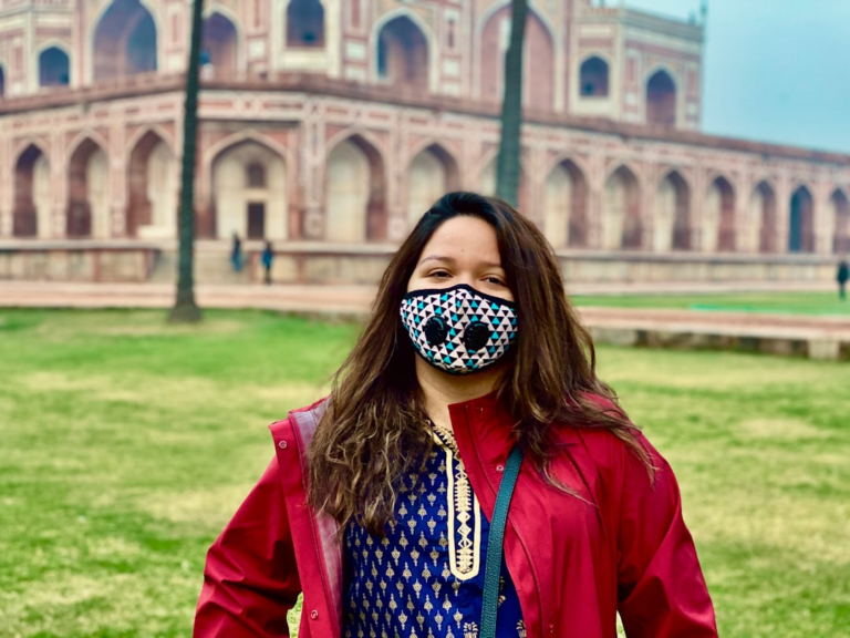 A woman wearing a distinctive protective face mask and red jacket stands on a lush green lawn before the majestic, red-stone Humayun's Tomb, evoking a sense of modern health awareness amidst timeless beauty.