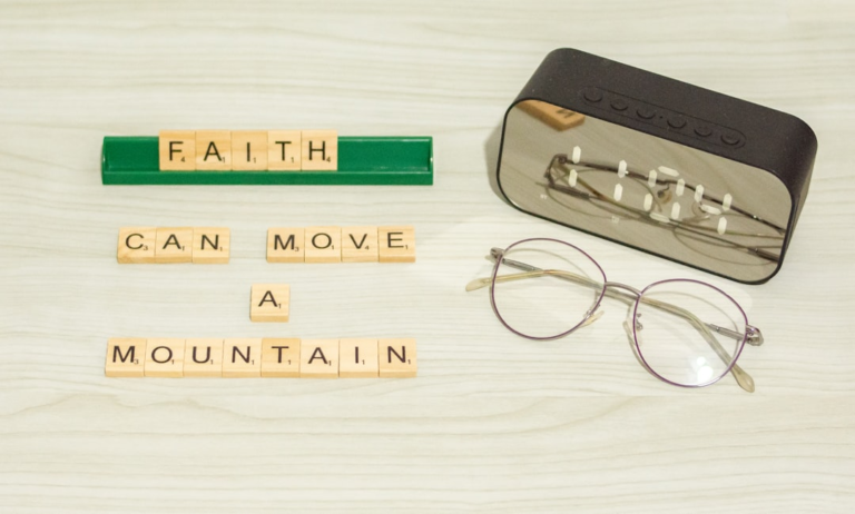 Scrabble tiles spelling Faith Can Move a Mountain are arranged on a light wood surface alongside modern eyeglasses and a digital clock, illustrating a calm, focused space ideal for clear vision and optimized lighting.