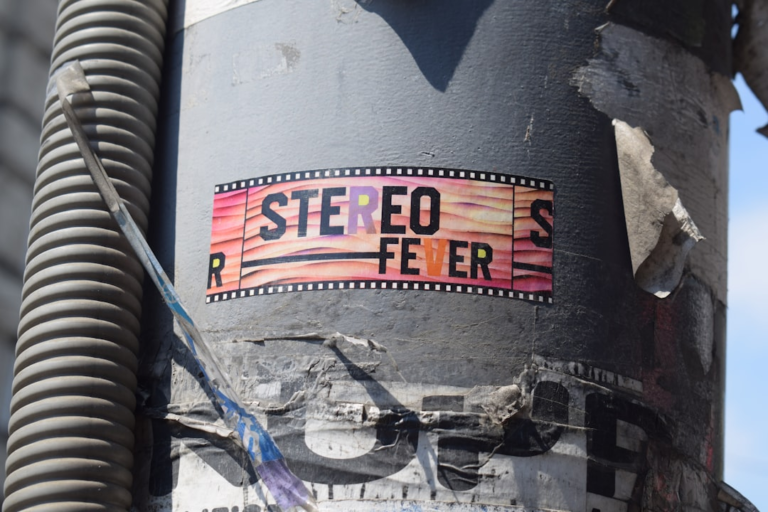 A colorful STEREO FEVER sticker with a wavy, film-strip design on a textured grey utility pole, offering a dynamic visual target for binocular vision exercises.