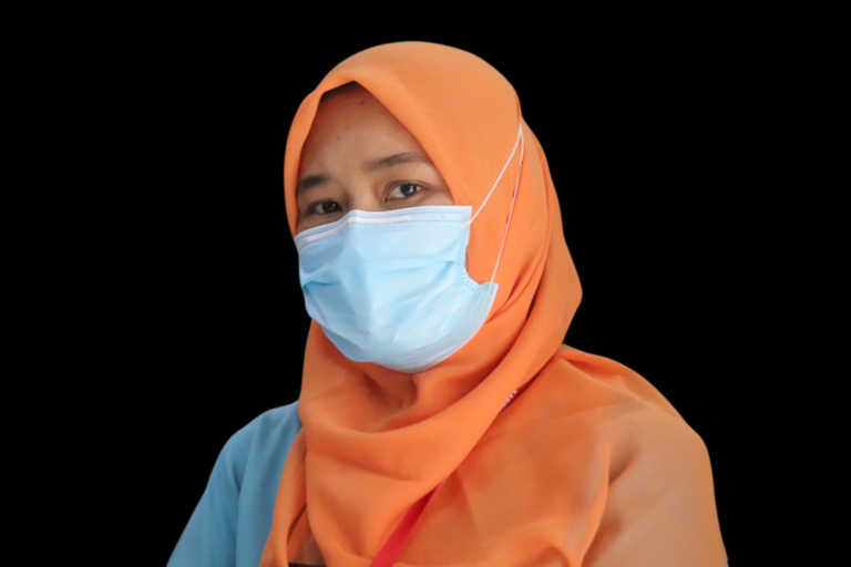 A person wearing an orange hijab and a light blue medical mask looks directly forward, their visible eyes clear and attentive against a dark background, embodying a focus on health and well-being.