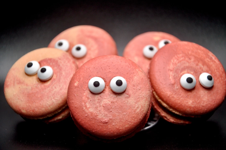Group of whimsical, reddish-pink macarons with googly eyes arranged on a dark background, humorously representing the importance of diet for healthy vision and combating Age-related Macular Degeneration (AMD).