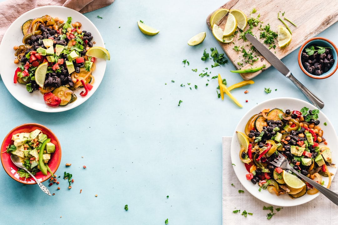 Two vibrant plates of black bean and colorful vegetable medley, garnished with fresh avocado and lime, sit alongside preparation ingredients on a light blue surface, representing a nutritious, plant-based diet beneficial for macular degeneration.