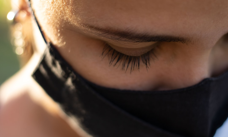 Close-up of a person's eye gently closed with long eyelashes, wearing a black face mask, and bathed in soft sunlight, conveying a sense of calm and relief from digital eye strain.