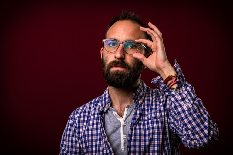 Bearded man in a plaid shirt adjusts his blue light blocking glasses, featuring a prominent blue reflection on the lenses, as he looks directly at the viewer against a dark red background, representing digital eye protection and visual clarity.