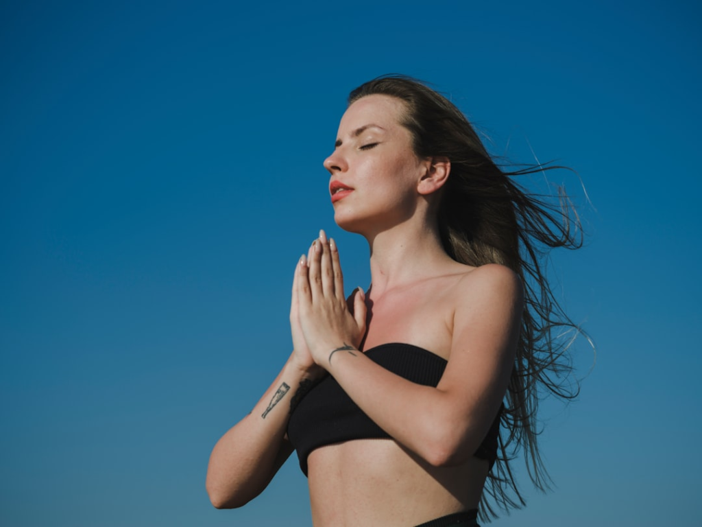 Serene young woman with eyes closed and hands clasped in a calming gesture against a brilliant blue sky, representing mindfulness and stress resilience.