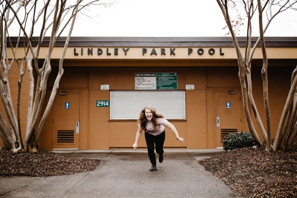 A spirited young woman with an excited expression vigorously runs forward from the Lindley Park Pool building, flanked by bare trees and a mulched path, symbolizing the high energy and metabolic boost from exercise.