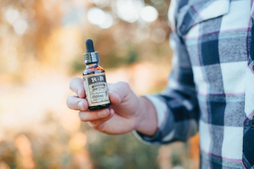 A hand, belonging to a person wearing a plaid shirt, holds a small brown dropper bottle of R+R Medicinal Hemp Extract 2500mg against a soft, sun-dappled outdoor background.