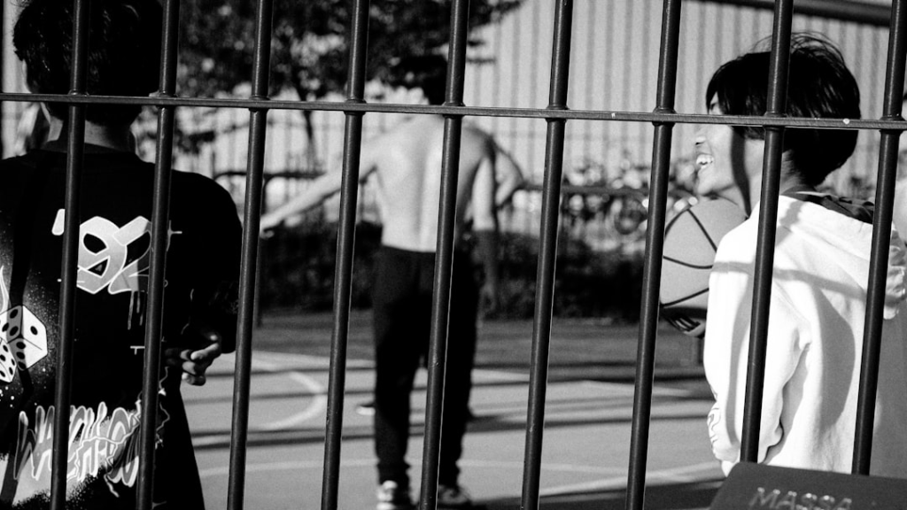 Black and white scene of active youth playing basketball on an outdoor court, with one person smiling brightly while holding a ball, viewed through a metal fence, symbolizing vitality and energy.