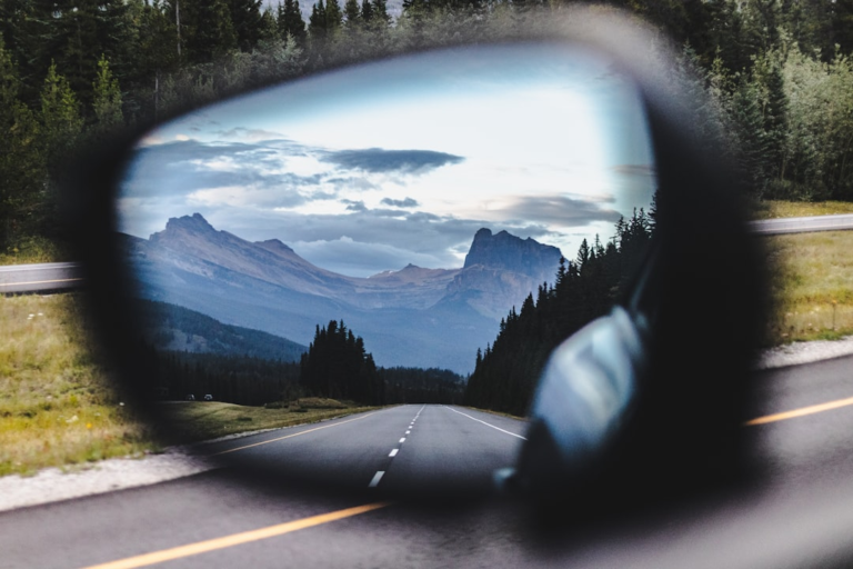 A car's side mirror clearly reflects a long, winding highway stretching toward majestic mountain peaks and dense pine forests under a vast, cloudy sky.