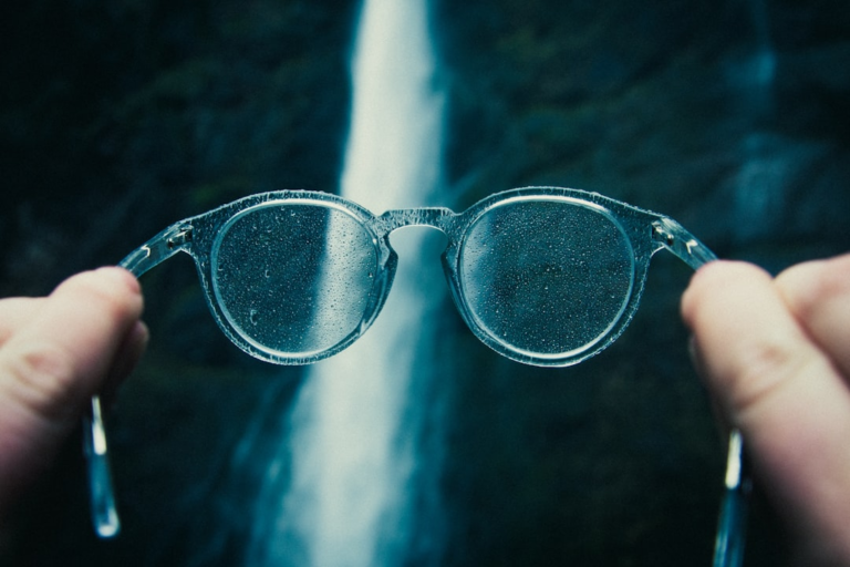 Hands hold water-splashed clear eyeglasses in front of a blurred, majestic waterfall, symbolizing the journey towards natural vision improvement and enhanced clarity.