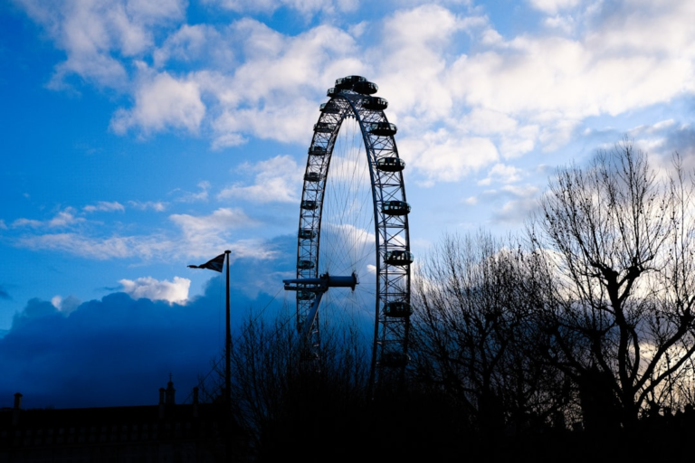 A towering Ferris wheel and bare trees are silhouetted against a vibrant blue sky with dramatic clouds, highlighting the powerful presence of light and an iconic urban structure.