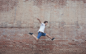 Energetic young man in a white shirt and denim shorts joyfully leaps mid-air against a weathered brick wall, embodying vitality and optimal metabolic health.