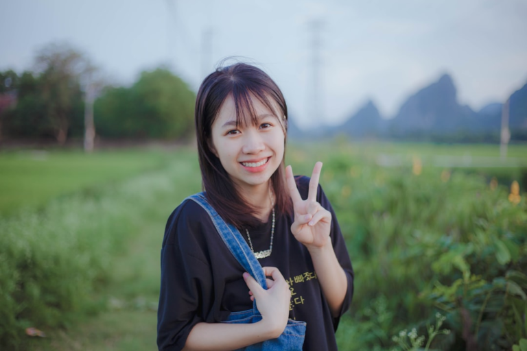 Cheerful young woman with bangs smiles and makes a peace sign in a lush green field, with distant mountains and trees in the natural background, conveying a sense of vitality and well-being.