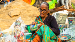 Elderly woman with glasses intently hand-painting a green and yellow traditional pot, surrounded by various detailed folk art crafts at an outdoor market stall.