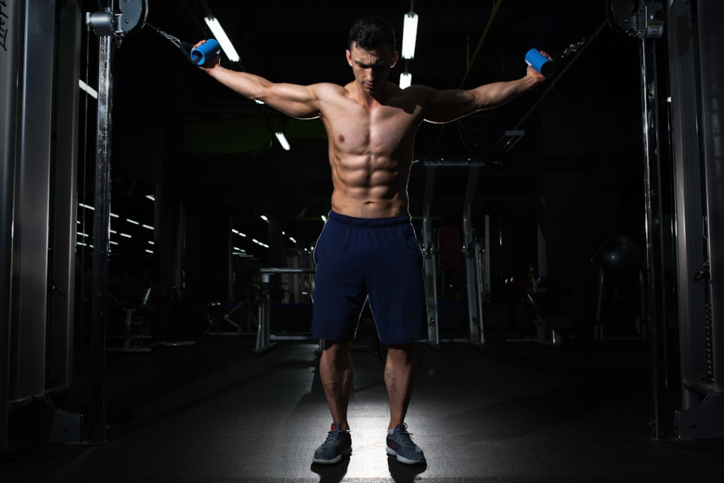 Muscular man with prominent abs performing a cable fly exercise in a darkly lit gym, embodying focused strength training for enhanced fat loss and improved insulin sensitivity.