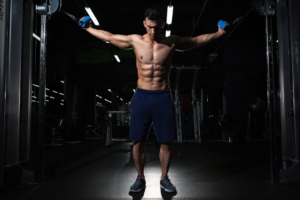 Muscular man with prominent abs performing a cable fly exercise in a darkly lit gym, embodying focused strength training for enhanced fat loss and improved insulin sensitivity.