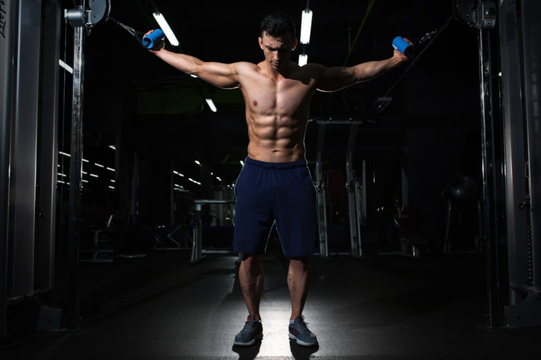 Muscular man with prominent abs performing a cable fly exercise in a darkly lit gym, embodying focused strength training for enhanced fat loss and improved insulin sensitivity.
