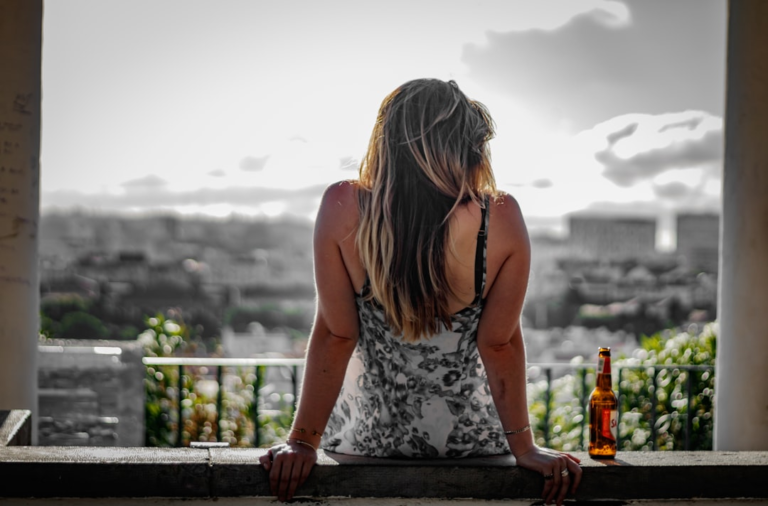 A woman with long hair sits illuminated by natural light, looking out over a desaturated city skyline with a beer bottle beside her, embodying a moment of relaxation and unwinding for recovery.