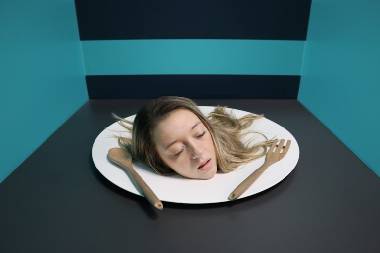 Surreal optical illusion featuring a young woman's blonde head on a large white dinner plate with wooden utensils, set in a teal and grey room, symbolizing the concept of bioavailability and absorbing vital nutrients for vision health.