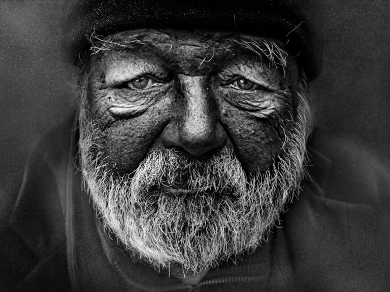 A high-contrast black and white portrait of an elderly man with a deeply textured, wrinkled face and a prominent white beard, embodying the visible effects of aging and the concept of human longevity.