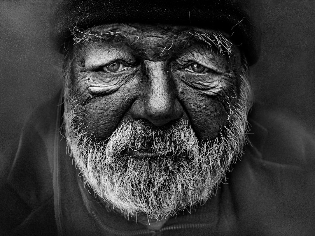 A high-contrast black and white portrait of an elderly man with a deeply textured, wrinkled face and a prominent white beard, embodying the visible effects of aging and the concept of human longevity.