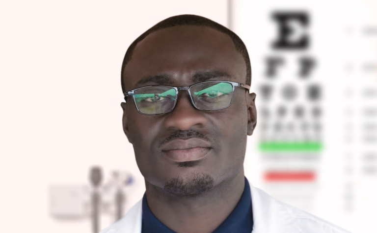 A professional Black man in glasses and a white lab coat stands in an ophthalmology clinic, with a blurred Snellen eye chart and medical equipment visible in the background, conveying a focus on vision health.