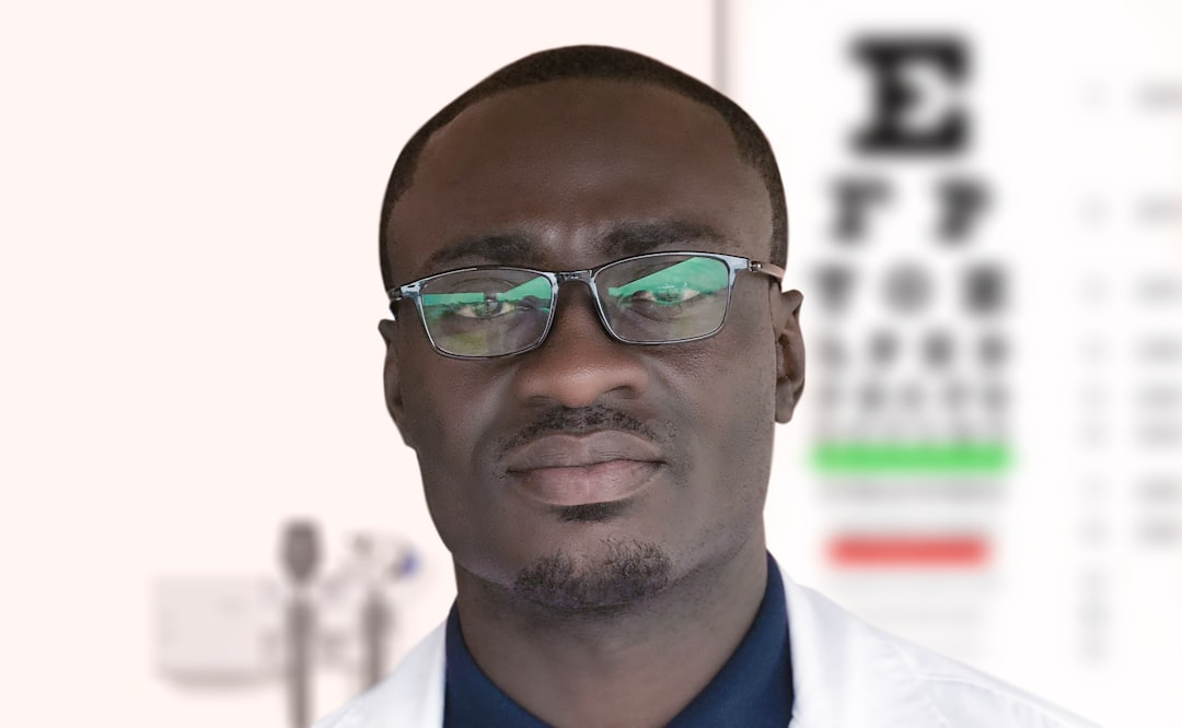 A professional Black man in glasses and a white lab coat stands in an ophthalmology clinic, with a blurred Snellen eye chart and medical equipment visible in the background, conveying a focus on vision health.