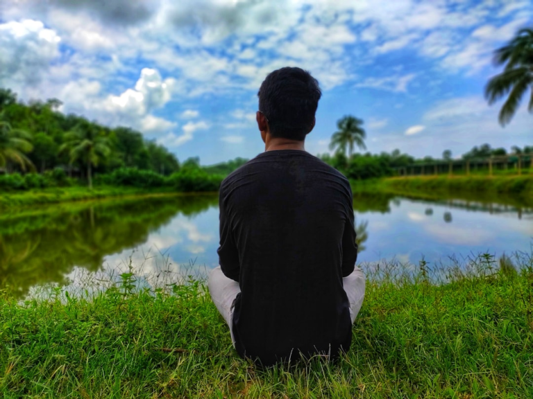 Man calmly sits by a tranquil lake surrounded by lush green nature under a partly cloudy sky, a serene scene promoting mental clarity and natural well-being.