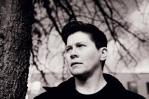 A person with short dark hair gazes reflectively upwards in a stark black and white outdoor scene featuring bare tree branches and a tree trunk, suggesting thoughtful consideration of natural well-being strategies.